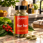A bottle of Jahn Herbs 'for her' dietary supplement capsules sits on a polished marble kitchen counter, surrounded by fresh green leaves, red berries, and scattered capsules.