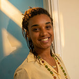 A smiling woman with braided hair stands indoors near a window, wearing a light beige short-sleeve collared shirt, a green beaded necklace, and a tan skirt while holding papers.